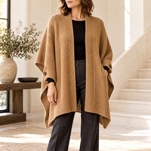 May include: A tan knit open-front poncho featuring a textured vertical ribbed pattern, a wide collar, and dark brown buttons on the sides. The garment is worn over a black long-sleeve top and dark gray trousers, creating a layered look.