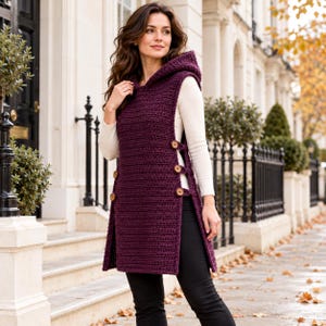 May include: A deep purple sleeveless hooded tunic featuring a textured crochet knit pattern. The sides of the garment are open and secured with three brown wooden buttons on each side, layered over a long-sleeved white shirt and black pants.