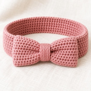 Può includere: Una fascia all'uncinetto color rosa polvere con un fiocco centrale prominente. La fascia è realizzata con un motivo a maglia denso e uniforme, e il fiocco è fissato con un centro in filo strettamente avvolto, creando un aspetto coeso e strutturato.