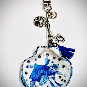 May include: A decorative keyring featuring a white seashell painted with a blue sea turtle and small blue beads. The shell hangs from a silver-toned metal chain with a lobster clasp, adorned with a small silver shell charm, a tree charm, a turtle charm, and a blue tassel.