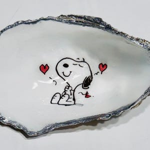 May include: A white oyster shell dish with a silver-colored edge. The dish features a hand-drawn illustration of Snoopy with two red hearts. The cartoon character is smiling and appears to be hugging himself.