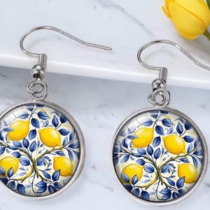 May include: A pair of round drop earrings featuring a silver-toned metal frame and a glass cabochon insert. The design displays a repeating pattern of bright yellow lemons and deep blue leaves against a light, off-white background.
