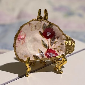 May include: A decorative, shell-shaped dish with a floral design and gold trim, displayed on a gold-colored stand. The dish features red flowers, pearls, and a butterfly. The stand is approximately 4 inches tall.