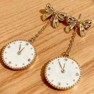 May include: A pair of gold-colored earrings featuring a bow design at the top and a clock face pendant. The clock face is white with gold accents and small crystals. The earrings are on a wooden surface.