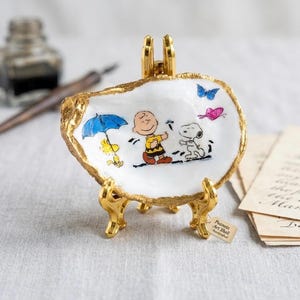 May include: A decorative, white, shell-shaped dish with a gold-painted rim, supported by a gold stand. The dish features a colorful illustration of Charlie Brown, Snoopy, and Woodstock from the Peanuts comic strip. A small tag reads "Peanuts Art Shell Handcrafted."