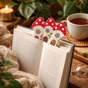 Könnte beinhalten: Drei pilzförmige Lesezeichen mit roten und weißen Kappen und cremefarbenen Körpern stecken in einem offenen Buch. Jedes Lesezeichen zeigt ein kleines Hausdesign mit Tür, Fenster und floralen Akzenten. Eine Tasse Tee, eine brennende Kerze und eine Brille sind ebenfalls zu sehen.
