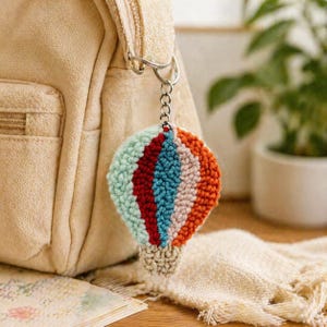 Puede incluir: Un llavero de globo aerostático hecho a mano. El llavero está hecho de hilo texturizado en tonos de azul claro, rojo, turquesa, rosa y naranja. Está unido a una cadena de metal plateado y a una mochila beige.