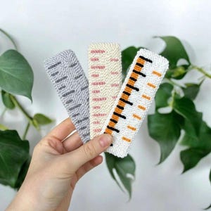 May include: Three handmade fabric bookmarks in a hand. The bookmarks feature textured designs in gray, cream with pink, and white with orange and black. The bookmarks are rectangular and appear to be made of woven fabric.