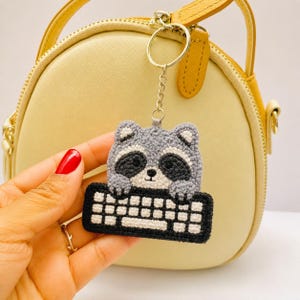 Può includere: Portachiavi a forma di procione grigio e nero con design a tastiera. Il portachiavi è realizzato in materiale all'uncinetto e ha una catena e un anello color argento. Lo sfondo include una borsa gialla con un cinturino giallo.