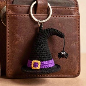 May include: A crocheted black witch hat keychain featuring a purple band with a yellow buckle and a small black spider dangling from the tip. The hat is attached to a silver metal keyring, showcasing detailed yarn stitching and a textured finish.