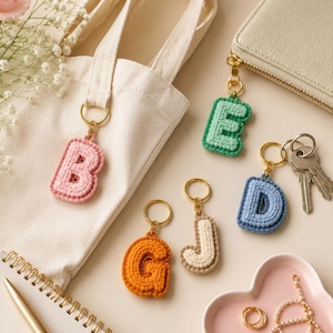 以下が含まれることがあります： カラフルな手編みの文字キーホルダーのコレクション。文字はB、E、D、G、Jで、それぞれピンク、グリーン、ブルー、オレンジ、クリーム色です。キーホルダーはゴールドカラーのリングに付けられ、財布、鍵、トートバッグと一緒に展示されています。