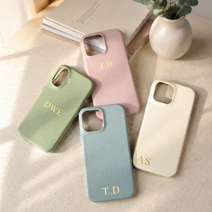 Puede incluir: Cuatro fundas de teléfono personalizadas en colores pastel: verde claro, rosa, azul y crema. Cada funda tiene un monograma dorado: "DWE", "LB", "T.D" y "AS". Las fundas están dispuestas sobre una superficie clara.