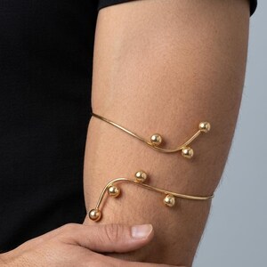 Peut inclure: Un bracelet de bras en métal doré au design à double bande qui s'enroule autour du haut du bras. La pièce est ornée de petites perles sphériques lisses fixées à intervalles le long de la structure en fil fin et incurvé du bijou.