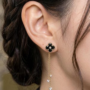 Peut inclure: Une boucle d'oreille en or avec un motif de trèfle à quatre feuilles composé de pétales en onyx noir et d'une pierre centrale transparente. Deux fines chaînes en or pendent du trèfle, ornées chacune d'une petite pierre transparente étincelante.