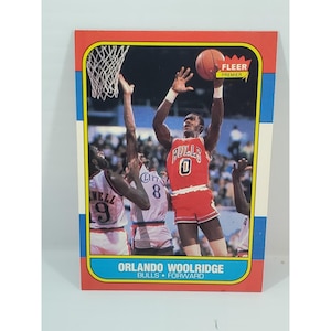 May include: A vintage Orlando Woolridge basketball card from the Fleer Premier series. The card features a player in a red Bulls uniform, mid-air, shooting a basketball. The card has a red border with blue and white stripes and the text "Orlando Woolridge Bulls Forward".