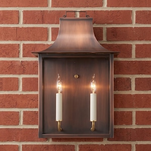 Può includere: Una lanterna da parete rettangolare per esterni con finitura in bronzo scuro e una copertura superiore curva. L'apparecchio presenta due luci verticali a candela con manicotti bianchi e lampadine a fiamma su una piastra posteriore piatta.
