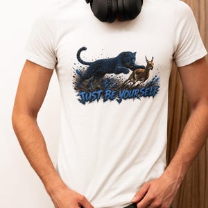 以下が含まれることがあります： 白い半袖Tシャツ。黒いヒョウが茶色のガゼルを追いかける様子が、青と茶色のテクスチャのある地面と共に描かれています。動物の下には「JUST BE YOURSELF」という文字が青いフォントでプリントされています。