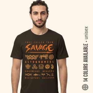 May include: A dark brown short-sleeved t-shirt featuring orange graphic text and symbols. The text reads: THEY CALLED THEM SAVAGE, ASTRONOMERS, ENGINEERS, HEALERS, NAVIGATORS, BUILDERS. Various ancient-style icons are arranged between the words.