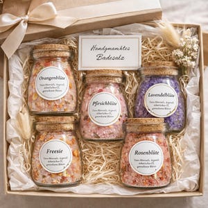 Könnte beinhalten: Eine Geschenkbox mit fünf Gläsern Badesalz mit Korkdeckeln. Die Gläser sind mit farbigen Salzen und getrockneten Blumen gefüllt. Die Etiketten tragen die Aufschrift "Orangenblüte", "Pfirsichblüte", "Lavendelblüte", "Freesie" und "Rosenblüte".