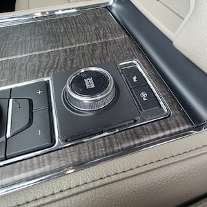 Puede incluir: Primer plano de la consola interior de un coche con un dial giratorio "DRIVE MODE" y botones de asistencia al aparcamiento. La consola tiene un acabado de veta de madera oscura, molduras plateadas y cuero beige.