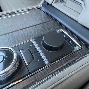 May include: Close-up of a car's interior console featuring a dark wood grain finish. The console includes a black control knob, buttons, and a silver-trimmed dial. The surrounding surfaces are light gray leather.