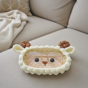 May include: A cream-colored, crocheted sheep-shaped glasses holder with brown ears and a brown nose. The holder contains a pair of gold-rimmed glasses. The holder is sitting on a beige couch.