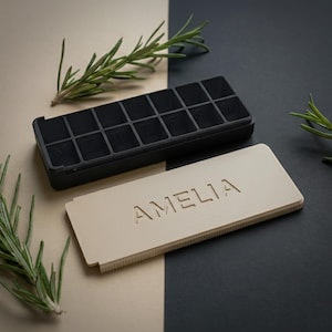 Puede incluir: Un pastillero negro con una tapa beige que dice "AMELIA". El organizador tiene 14 compartimentos. Ramas de romero se colocan alrededor del organizador sobre un fondo bicolor beige y negro.