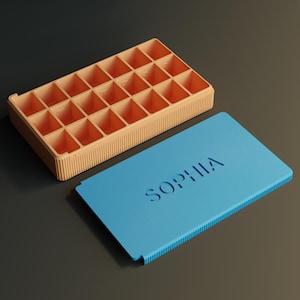 Op de afbeelding: Een open perzik-kleurige organizer met 24 vierkante compartimenten, naast een lichtblauw deksel. Het deksel is voorzien van de naam "SOPHIA". De organizer en het deksel liggen op een donkergrijze ondergrond.