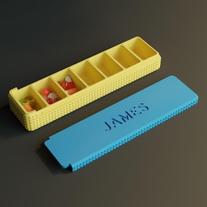 May include: A yellow rectangular pill organizer with seven compartments, some containing assorted colorful capsules. A separate blue lid with the name JAMES cut out in a bold font sits beside the organizer. Both pieces feature a textured exterior.