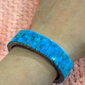 Peut inclure: Un bracelet extensible composé de segments rectangulaires en pierre turquoise bleu vif avec des inclusions naturelles de matrice sombre. Le bracelet est construit avec une bande métallique flexible qui lui permet de s'étendre autour du poignet.