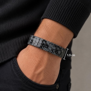 Può includere: Un bracciale composto da segmenti rettangolari in ossidiana fiocco di neve, caratterizzato da una base nera con motivi grigi simili a fiori. Il bracciale ha una larghezza di 1,5 cm, come indicato dal testo di misurazione visibile sul lato.
