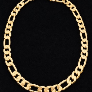 May include: A gold-toned Figaro chain necklace featuring a repeating pattern of three small circular links followed by one elongated oval link. The necklace is finished with a secure lobster claw clasp at the top, showcasing a polished metallic surface.