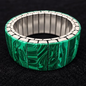 Op de afbeelding: Een rekbare armband met rechthoekige segmenten in een groen en zwart gemarmerd patroon. De buitenste segmenten zijn gerangschikt in een doorlopende cirkelvormige band, terwijl de binnenkant is gevoerd met gepolijste zilverkleurige metalen schakels.