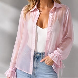 May include: A sheer, light pink button-down shirt with long sleeves and ruffled cuffs. The shirt is worn over a white tank top and blue jeans. The sleeves have a tie detail at the wrist.