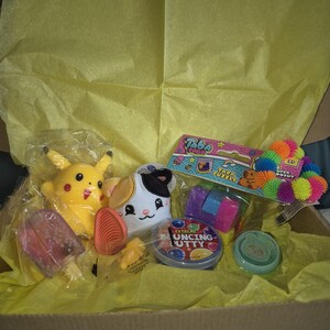 May include: A cardboard box containing a selection of toys and gifts, such as a yellow Pikachu plush, a cat plush, a popsicle, and a container of Extreme Bouncing Putty, all on yellow tissue paper.