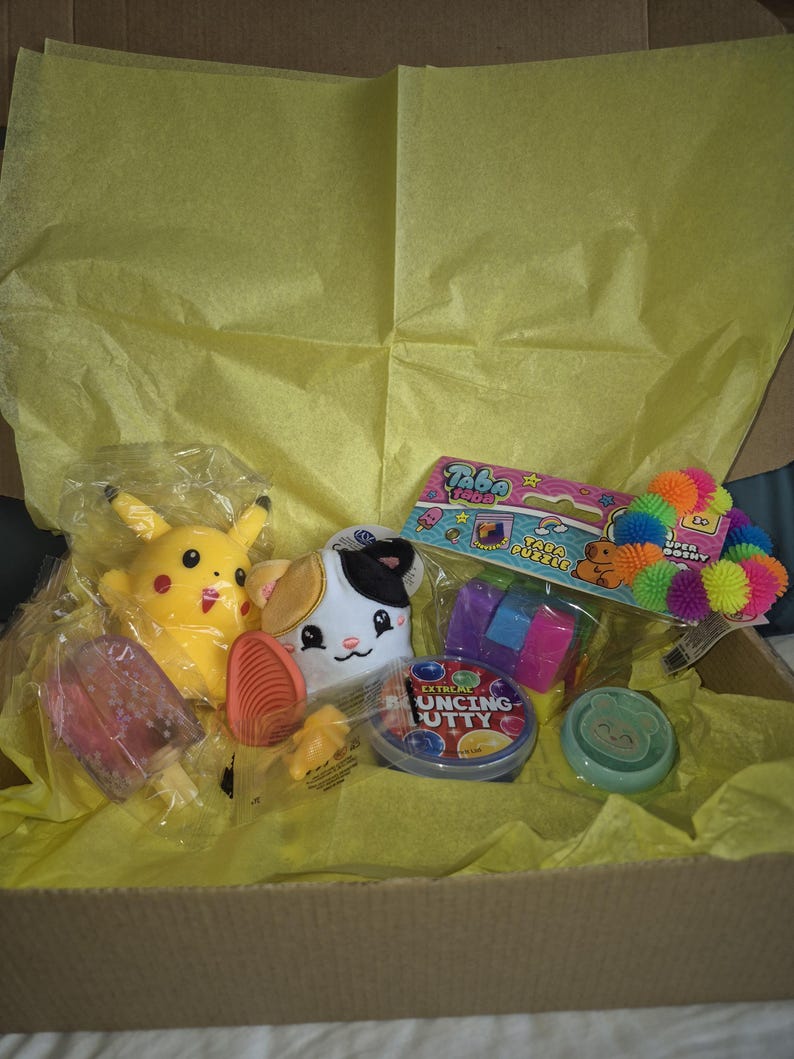 May include: A cardboard box containing a selection of toys and gifts, such as a yellow Pikachu plush, a cat plush, a popsicle, and a container of Extreme Bouncing Putty, all on yellow tissue paper.