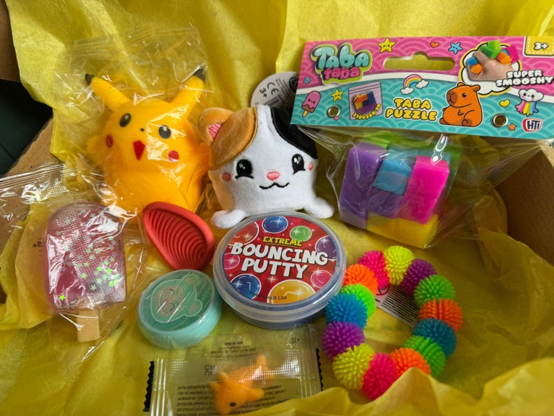 May include: A selection of toys in a box, featuring a yellow Pikachu plush, a white and calico cat plush, a Taba Puzzle, a container of Extreme Bouncing Putty, a rainbow spiky ball, and a popsicle-shaped toy, all in a yellow box.