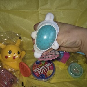 May include: A selection of colourful toys and novelty items, including a white plush toy with blue beads, a yellow Pikachu figure, and a container of bouncing putty. Other items include a red popsicle and a small orange figure.