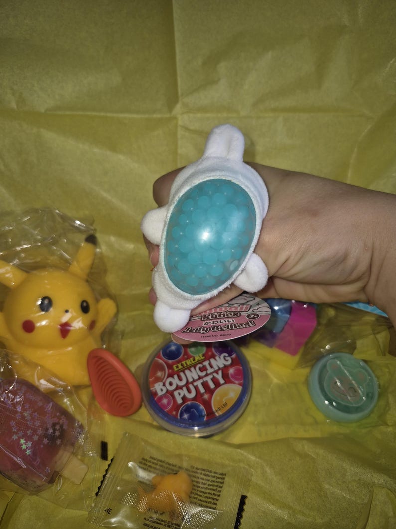 May include: A selection of colourful toys and novelty items, including a white plush toy with blue beads, a yellow Pikachu figure, and a container of bouncing putty. Other items include a red popsicle and a small orange figure.