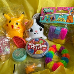 May include: A selection of toys and novelties in a box, featuring a yellow Pikachu plush, a white cat plush, a Taba Puzzle, a container of bouncy putty, and a rainbow spiky ball. The items are on yellow tissue paper.