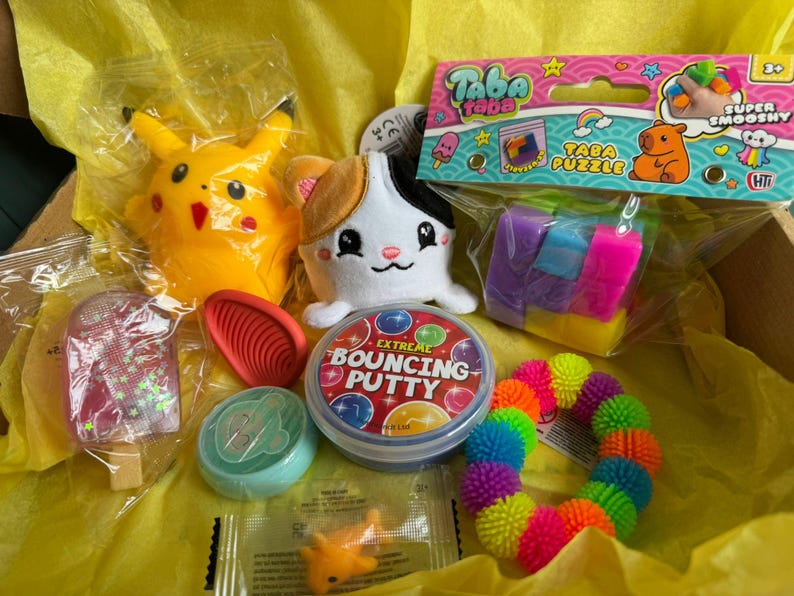 May include: A selection of toys and novelties in a box, featuring a yellow Pikachu plush, a white cat plush, a Taba Puzzle, a container of bouncy putty, and a rainbow spiky ball. The items are on yellow tissue paper.