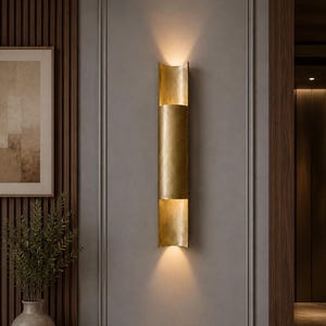 May include: A vertical wall sconce with a cylindrical brass finish, featuring an open-ended design that casts light both upward and downward. The fixture has a smooth, metallic surface and is mounted centrally on a flat, gray paneled wall surface.