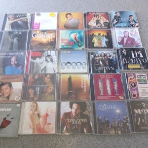 May include: A selection of music CDs in clear cases, showcasing a variety of album covers. The artwork includes portraits, band images, and abstract designs. The CDs are displayed on a neutral-toned surface.