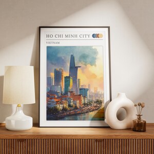 May include: Framed art print featuring a watercolor painting of Ho Chi Minh City, Vietnam. The artwork depicts a cityscape with skyscrapers, buildings, and a river, set against a colorful sunset. The print is displayed on a wooden surface.