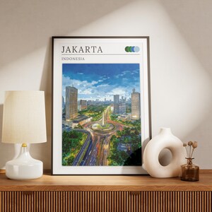 May include: A framed print of Jakarta, Indonesia, showing a cityscape under a blue sky. The print is displayed on a wooden cabinet, with a white lamp, a decorative vase, and a small glass bottle. The print has the word "Jakarta" at the top.