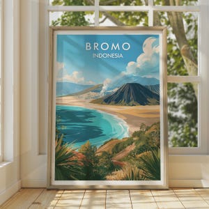 Op de afbeelding: Een ingelijste reisposter met een landschapsillustratie van de berg Bromo in Indonesië. Het kunstwerk toont een donkere vulkanische piek, een zandvlakte en een turquoise wateroppervlak onder een blauwe lucht met witte wolken. De tekst BROMO INDONESIA staat bovenaan in het wit gedrukt.