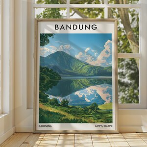 Puede incluir: Un póster de viaje enmarcado con una ilustración paisajística de Bandung, Indonesia. La obra muestra una gran montaña reflejada en un lago tranquilo, con colinas verdes en primer plano, un cielo azul con nubes blancas y el texto BANDUNG, INDONESIA, 6.92° S, 107.61° E.