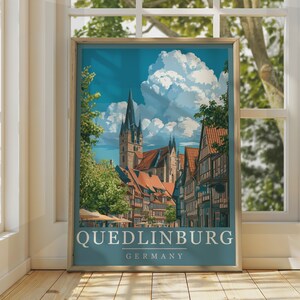 Puede incluir: Un póster de viaje enmarcado con una ilustración de Quedlinburg, Alemania, con casas de entramado de madera, una alta aguja de iglesia y grandes nubes blancas sobre un cielo azul. El texto Quedlinburg Germany está impreso en blanco abajo.