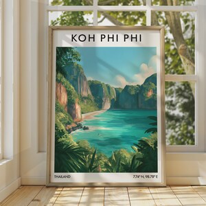 May include: A framed travel poster featuring a landscape illustration of Koh Phi Phi, Thailand. The artwork depicts a turquoise bay surrounded by lush green cliffs and a sandy beach, with the text "KOH PHI PHI", "THAILAND", and "7.74° N, 98.78° E".