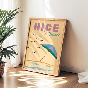 Könnte beinhalten: Ein gerahmter Kunstdruck mit einer Strandszene mit Sonnenschirm, Wellen und Möwen. Der Druck trägt das Wort "NICE" in Lila und "France" in Türkis. Der Rahmen ist hellbraun.